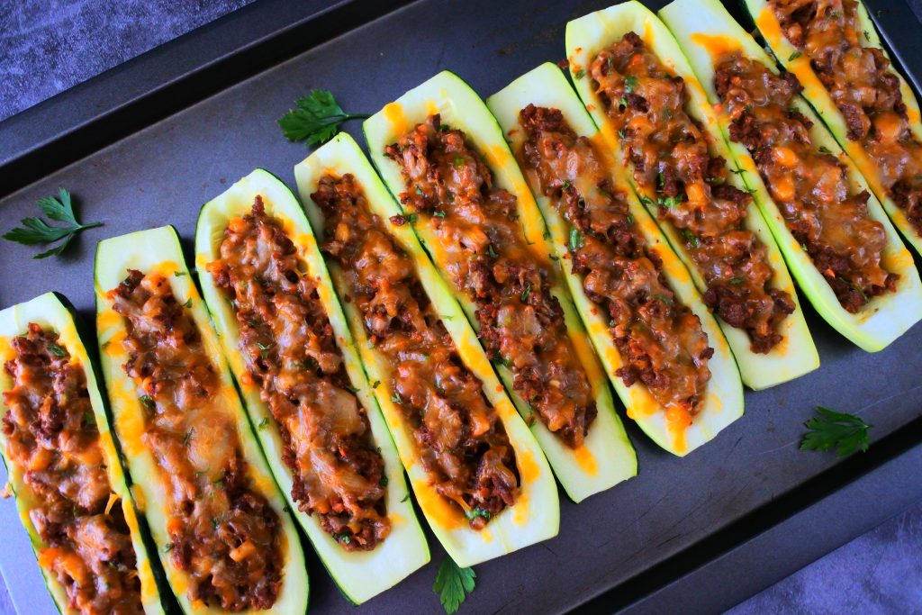 An angled overhead image of baked zucchini boats next to each other on a tray with leaves of fresh parsley around it