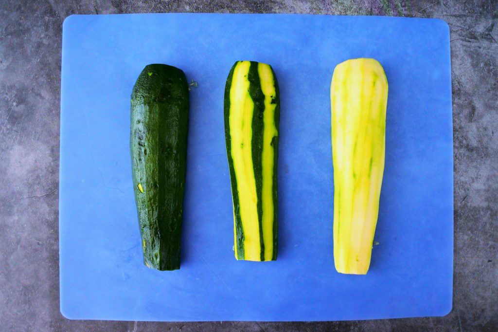 An overhead image of three zucchini halves, one with skin on, one with strips of skin removed and one peeled on a blue cutting mat