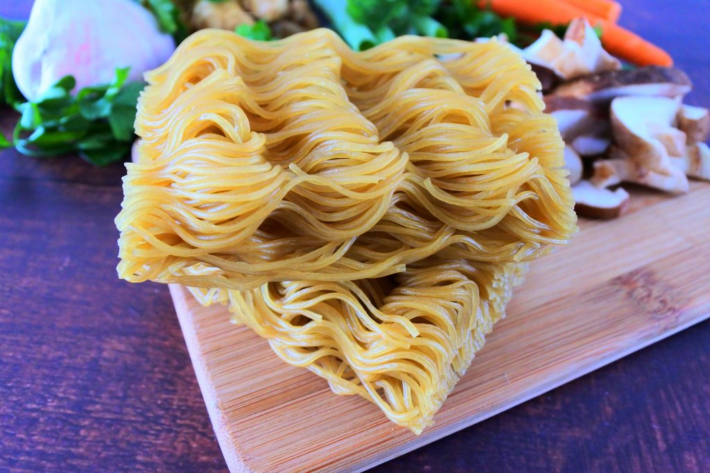 A close up image of two bricks of dry uncooked brown rice ramen noodles on a wood board with fresh vegetables in the background