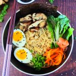 An overhead image of a plated bowl of chicken shoyu ramen topped with egg, mushrooms, bok choy, carrots, chicken, pickled ginger, chives and noodles. Surrounded by chopsticks, fresh herbs and a dish of black sesame seeds