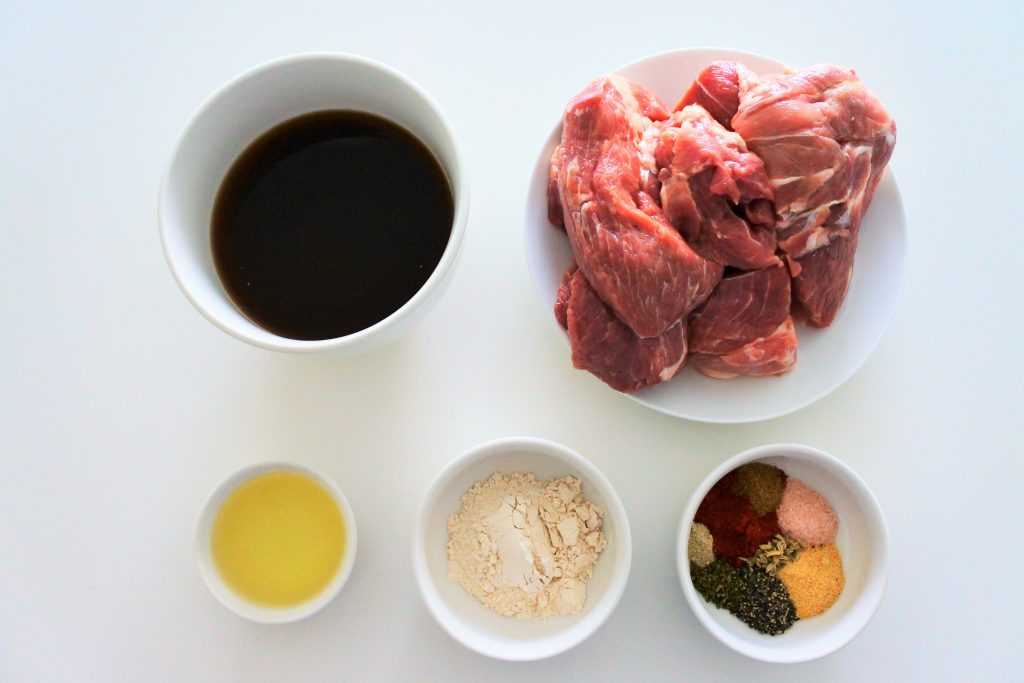 An overhead image of ingredients for oven roasted pork including beef broth, large pork chunks, oil, and herbs and spices for a rub