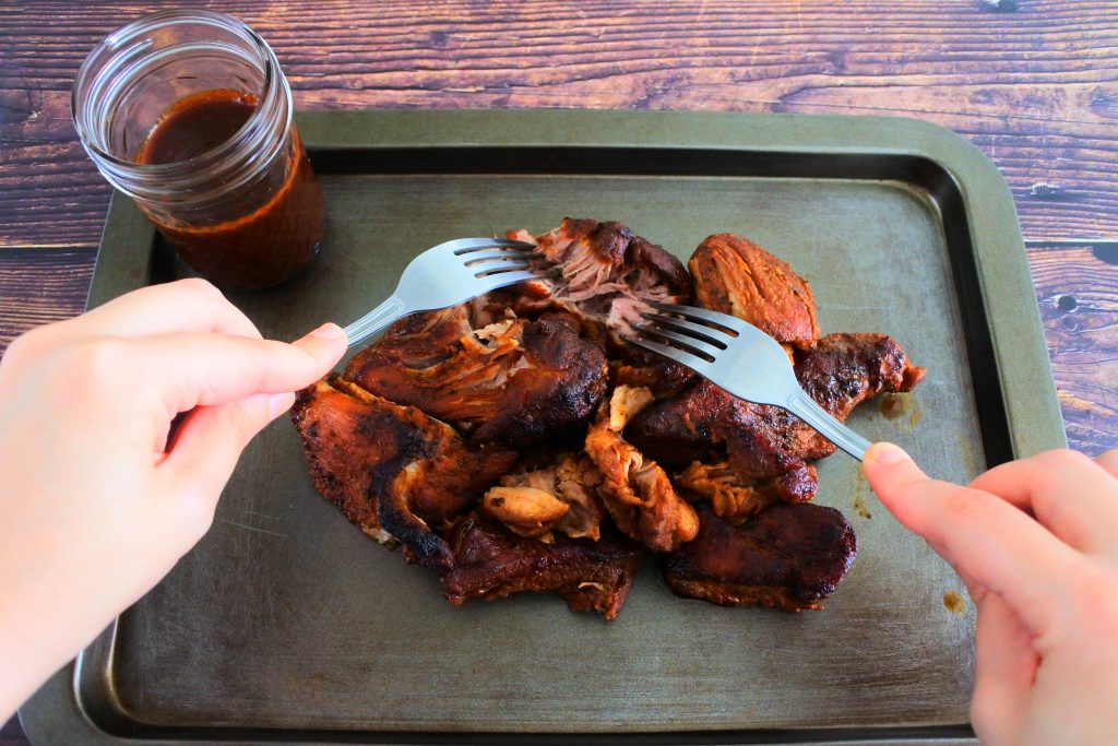 An angled image of slow oven roasted pork on a tray next to a jar of homemade bbq sauce being shredded with two forks