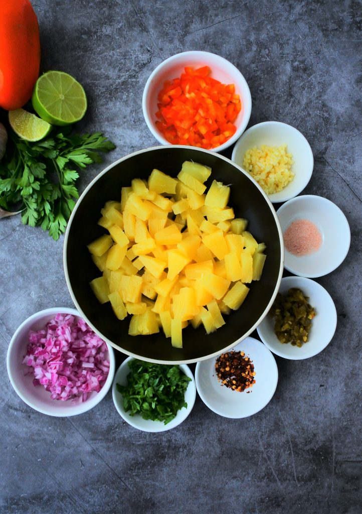 An overhead image of bowls of ingredients for a fresh pineapple salsa including chunks of pineapple and an assortment of herbs and spices