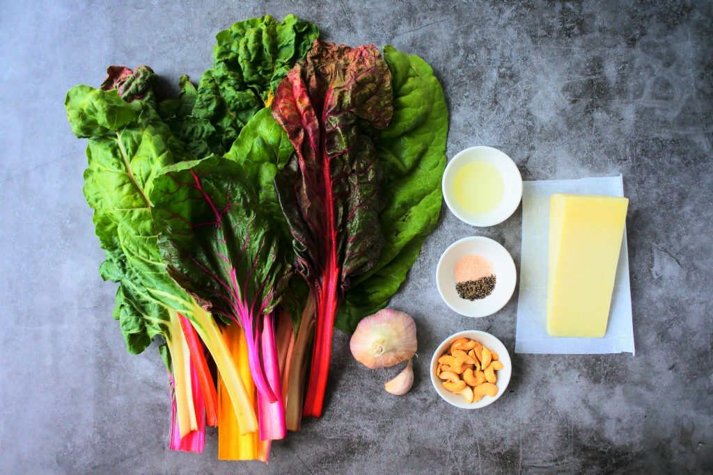 An overhead image of ingredients for sauteed chard including a bundle of rainbow chard, fresh garlic, oil, salt and pepper, cashews and a wedge of cheese
