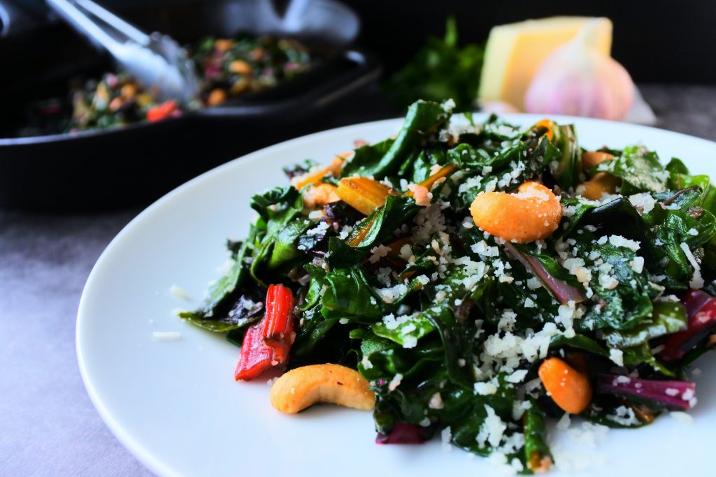 A head on close up image of sauteed rainbow chard on a plate dressed with cashews and shredded cheese with a head of garlic, a wedge of cheese and greens and a skillet with sauteed chard in the background
