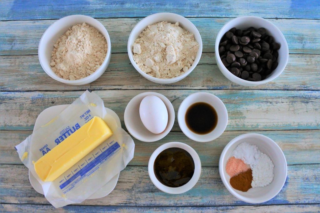 An overhead image of ingredients for a classic whole wheat cookie including whole wheat pastry flour, raw cane sugar, dark chocolate chips, butter, egg, vanilla extract, molasses, baking powder, baking soda, salt and cinnamon