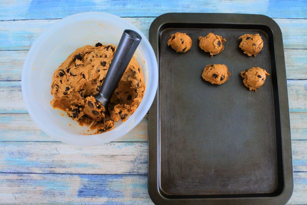An angled overhead image of a bowl of chilled chocolate chip cookie dough with a scooper in it and a tray with portions of dough on the side