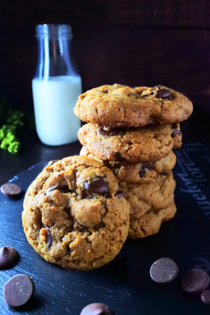 A stack of classic chocolate chip cookies with one cookie leaning against the side of the stack surrounded by chocolate chips with a bottle of milk in the background