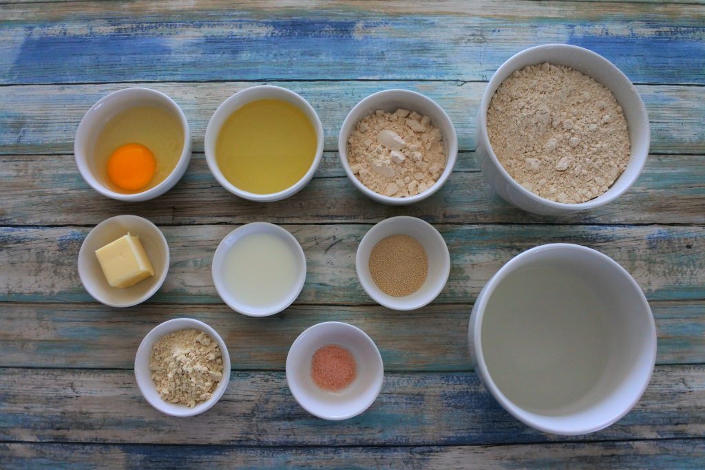 An overhead image of ingredients for one hour dinner rolls including whole wheat pastry flour, sugar, oil, egg, butter, milk, yeast, warm water, vital wheat gluten and salt