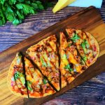 An overhead image of a BBQ chicken pizza flatbread on a board with fresh herbs and a block of cheese bordering the image