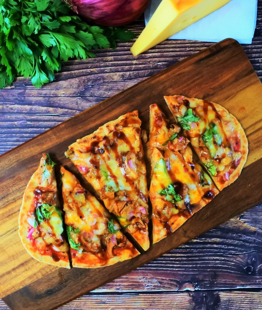 An overhead image of a BBQ chicken pizza flatbread on a board with fresh herbs and a block of cheese bordering the image