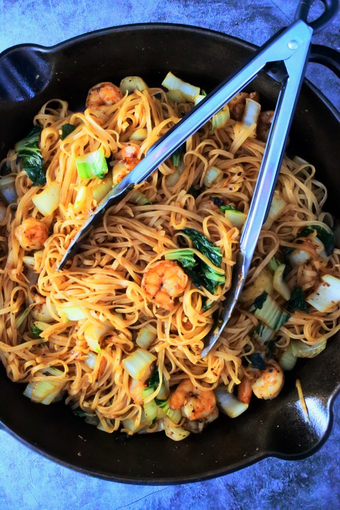 An overhead image of a skillet with shrimp noodle stir fry and a pair of tongs on top