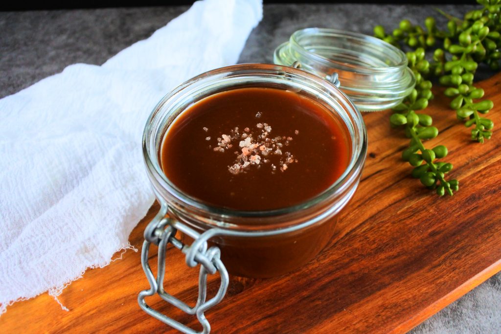 An angled image of a jar of salted caramel sauce topped with flakes of pink Himalayan salt on a wooden board.