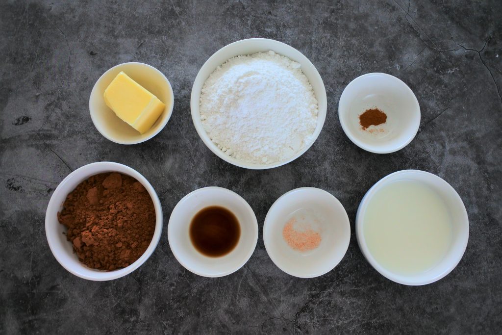 An overhead image of ingredients for homemade chocolate frosting including butter, finely blended cane sugar, cinnamon, cocoa powder, vanilla extract, salt and milk