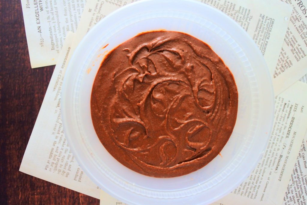 An overhead image of a bowl of chocolate frosting on scattered sheets of recipe pages