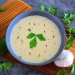 An angled image of a bowl of homemade Alfredo sauce in a stone bowl on a wooden plank surrounded by a small head of garlic, dried pasta and fresh parsley