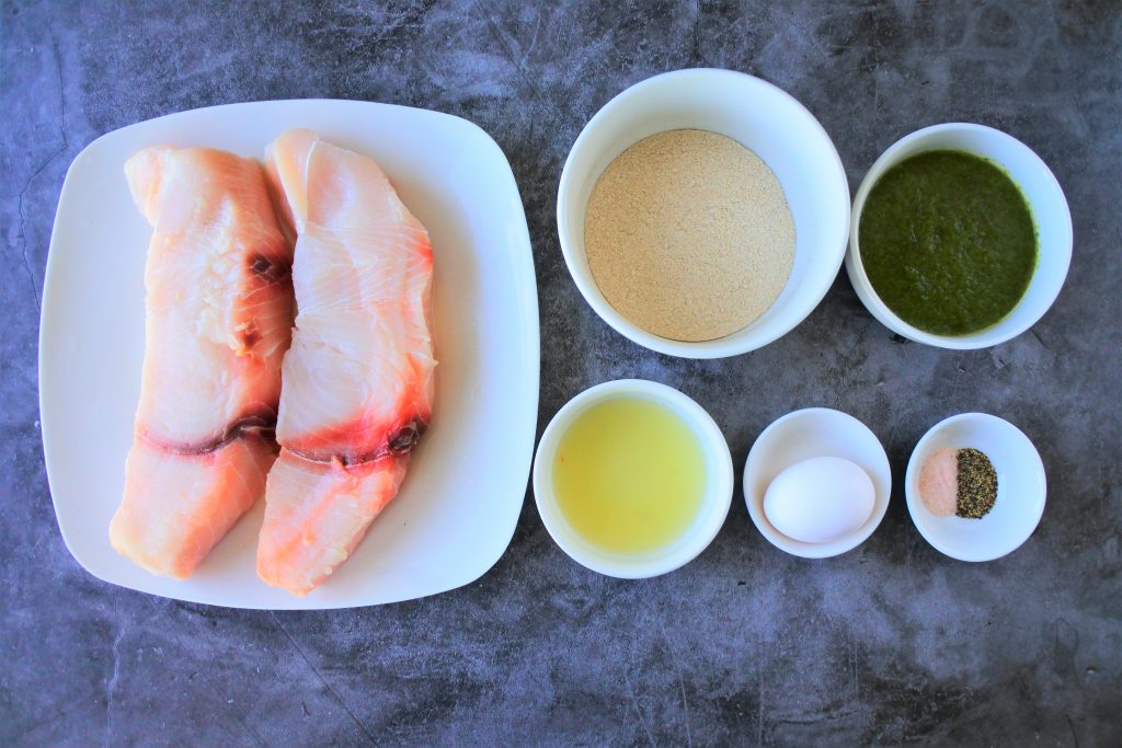 An overhead image of ingredients for Caribbean-style fried fish including fish (shark) fillets, whole wheat pastry flour, Green seasoning, lime juice, an egg and salt and pepper.