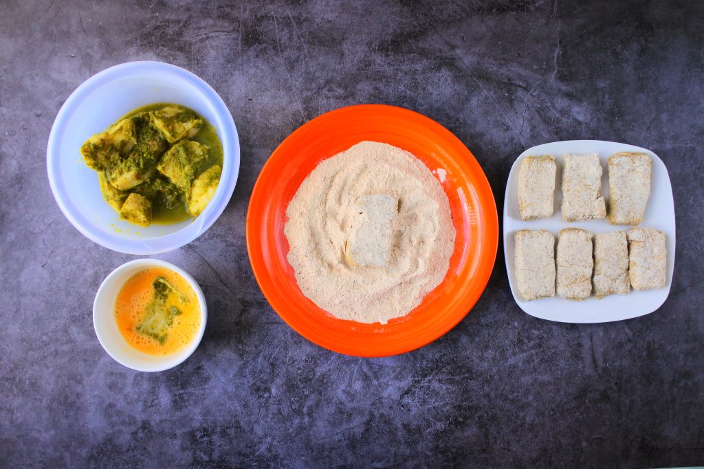 An overhead image of a fish breading line including seasoned fish fillets a bowl with a beaten egg, a plate with seasoned flour and a plate with breaded fillets