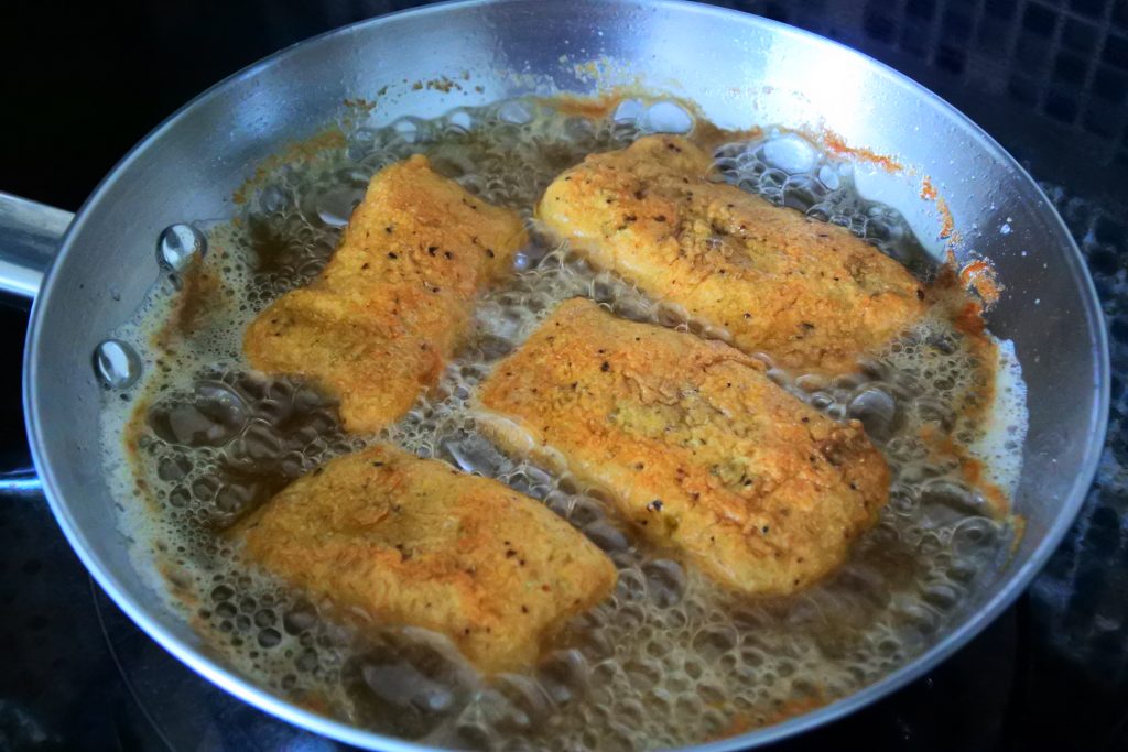 An angled image of a pan of breaded fish fillets being shallow fried in oil
