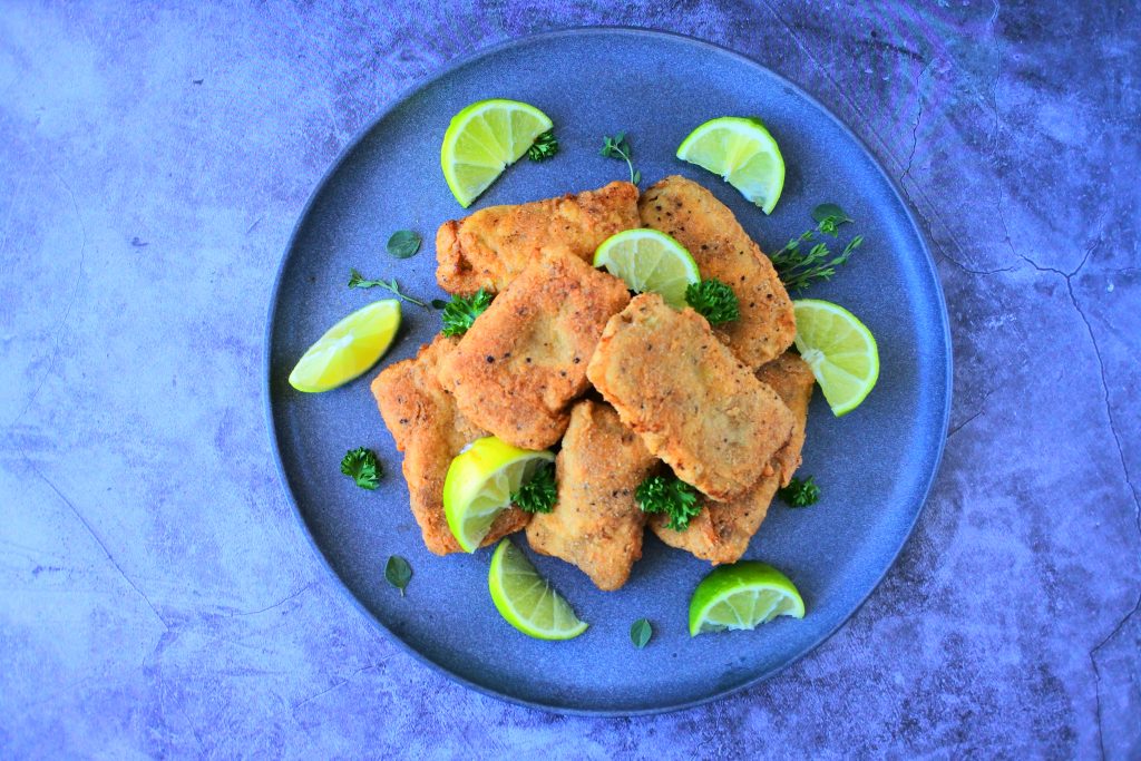 An overhead image of a plate of crispy fried fish fillets garnished with herbs and lime wedges