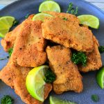 An angled image of a plate of crispy fried fish fillets garnished with herbs and lime wedges