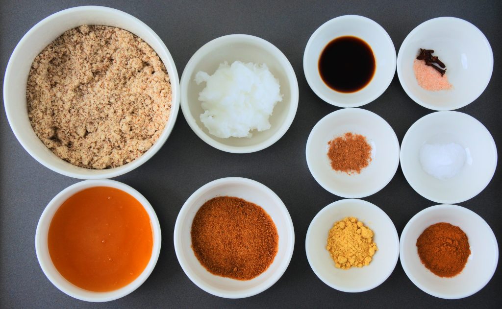 An overhead image of bowls of ingredients for chewy gingerbread cookies including: almond flour, coconut oil, raw honey, coconut sugar, vanilla extract, baking soda, salt, clove, nutmeg, ginger and cinnamon.