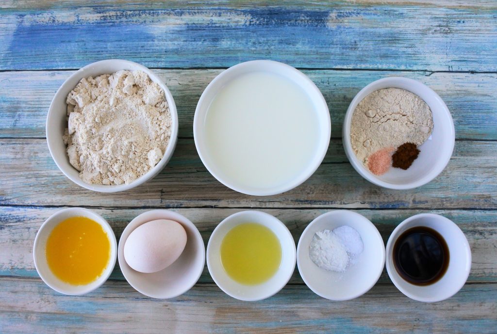 An overhead image of ingredients for whole wheat buttermilk pancakes including whole wheat pastry flour, milk, finely ground cane sugar, salt, cinnamon, vanilla extract, baking powder and baking soda, lemon juice, an egg, melted butter
