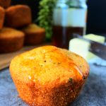 A close up image of a honey drizzled cornbread muffin with a jar of honey and a stack of muffins in the background