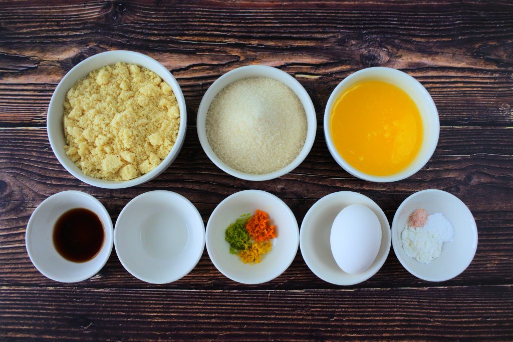 An overhead image of ingredients for a gluten free boterkoek / citrus butter cake including almond flour, sugar, butter, vanilla, orange extract, orange zest, egg, salt, baking powder and tapioca flour.