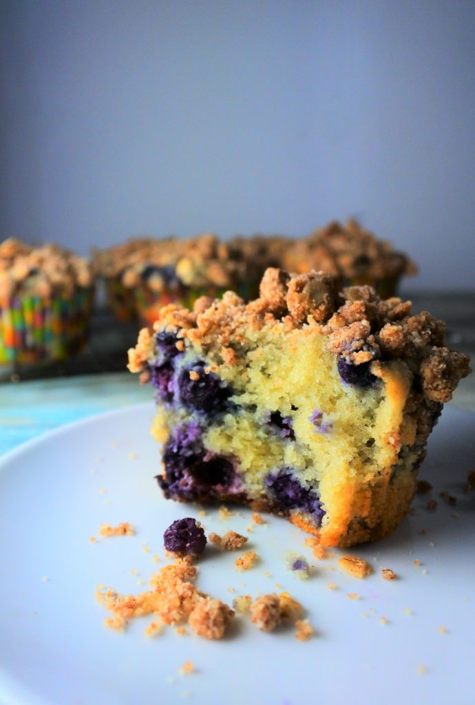 A head on image of a blueberry streusel muffin with a bite taken out of it on a plate with more muffins on a cooling rack in the background