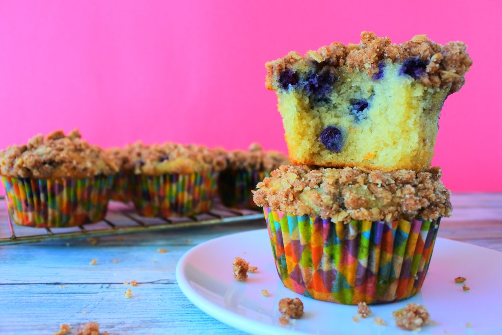 An image of two stacked blueberry streusel muffins with the top muffin missing a bite and a cooling rack with more muffins in the background