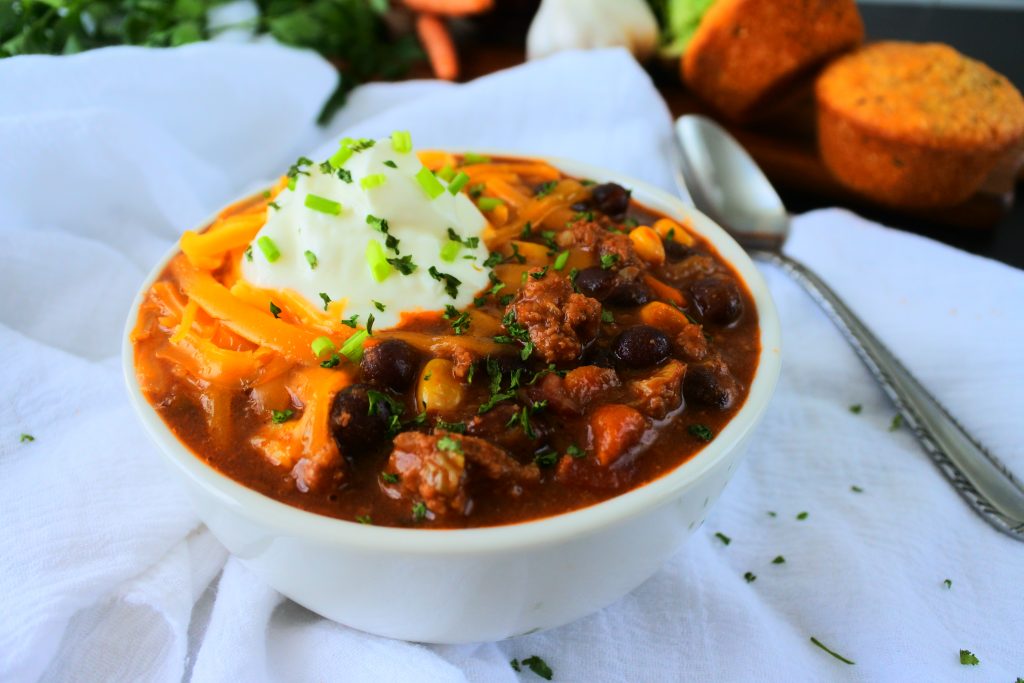 A head on close up image of a bowl of kitrusy's chili con carne topped with cheese, sour cream and chives with a spoon on the side