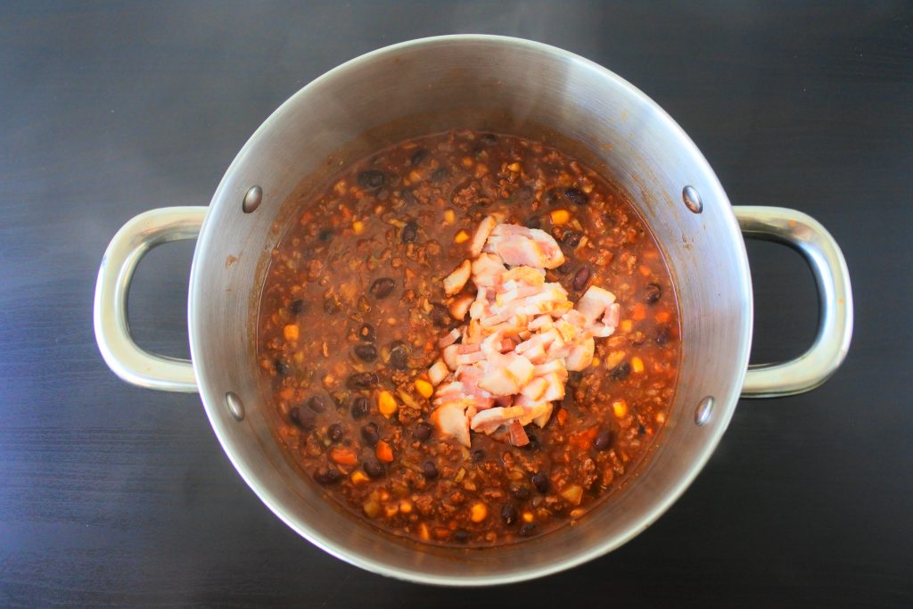 an overhead image of a pot of chili with pieces of bacon being added in