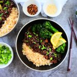 An overhead image of a ground beef teriyaki bowl served with steamed rice, sauteed broccolini and garnished with lemon wedges, chives and sesame seeds