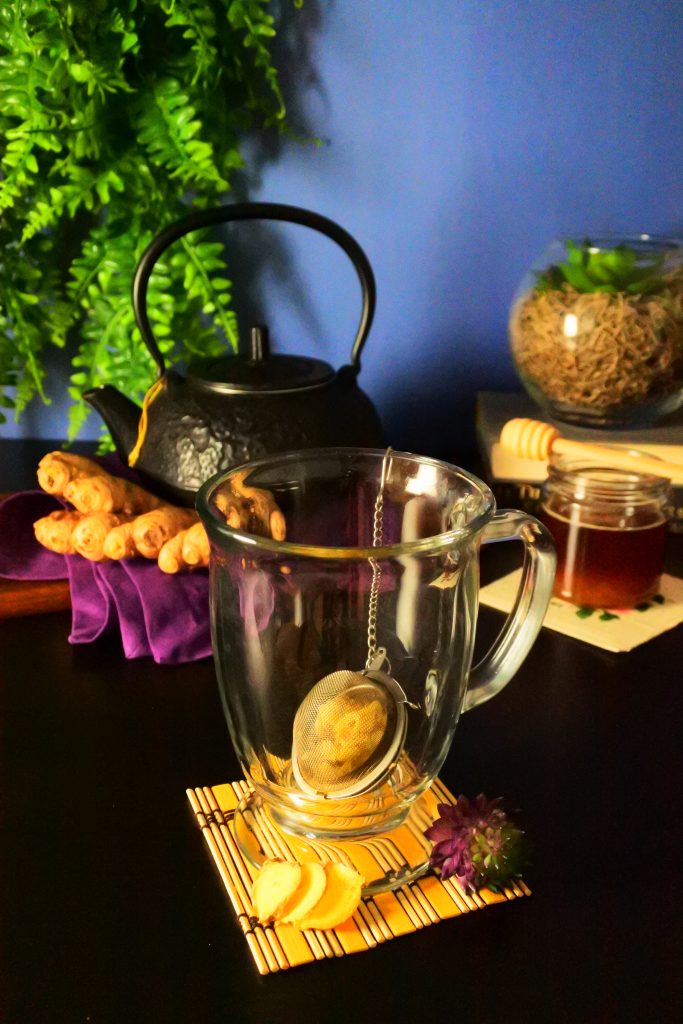 A head-on image of an glass mug with a ginger-filled tea strainer with a black kettle, fresh ginger, a small bottle of honey and plants in the background