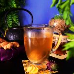A head-on image of a ginger-tea filled glass mug with a black kettle, fresh ginger, a small bottle of honey and plants in the background