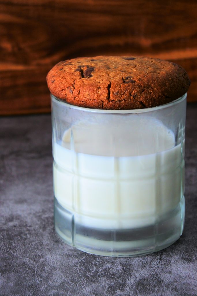 A head on image of a glass of milk with a warm, freshly baked gluten free chocolate chip cookie covering it