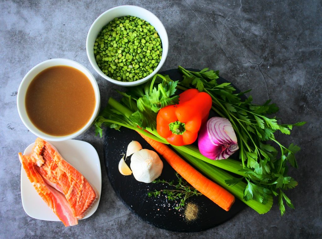 An overhead image of ingredients for a simple split pea and bacon soup including split peas, broth, bacon, and fresh herbs and vegetables