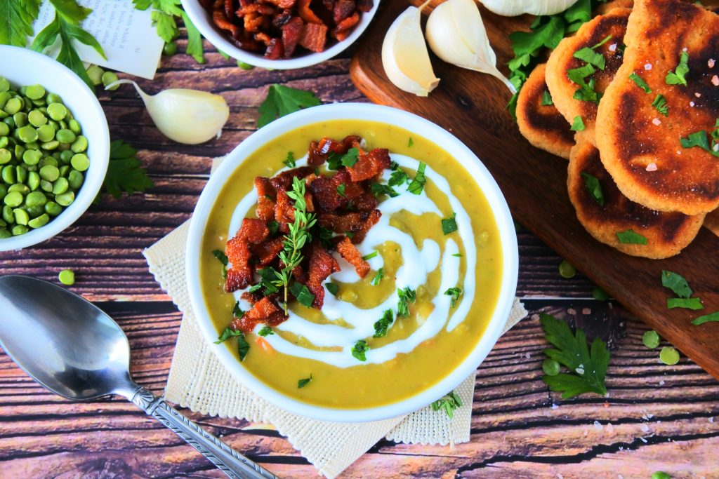 An overhead image of a bowl of green split pea soup topped with herbs and crispy bacon bits with other fresh ingredients surrounding it
