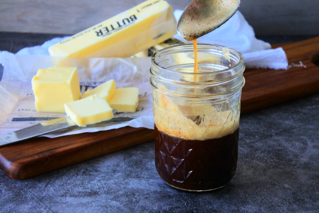 A head on image of browned butter in a mason jar with some being streamed from a spoon with sticks of butter on a wooden board in the background