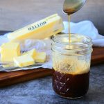 A head on image of browned butter in a mason jar with some being streamed from a spoon with sticks of butter on a wooden board in the background