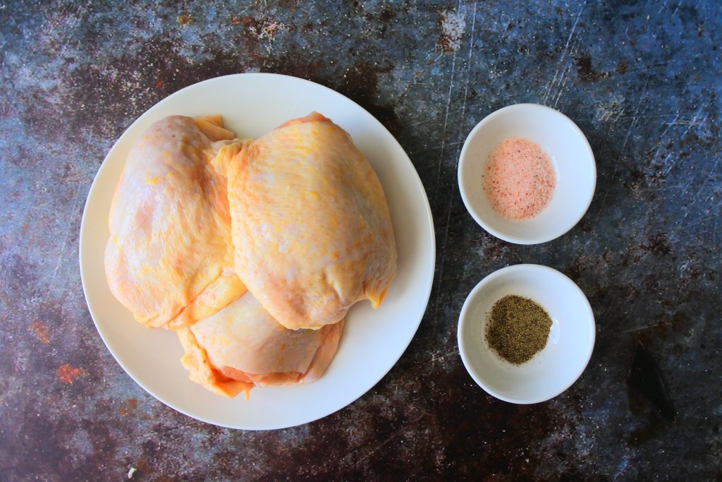 An overhead image of three skin on, bone in, chicken thighs next to two small dishes of salt and pepper