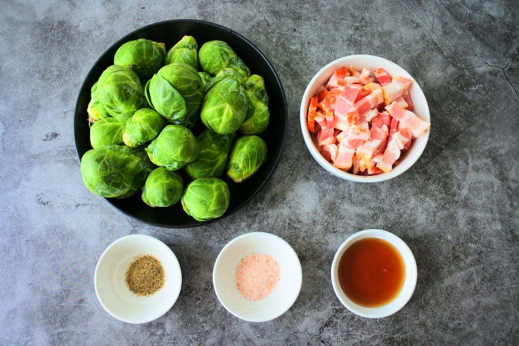 An overhead image of ingredients for maple bacon brussels sprouts including fresh brussels sprouts, diced thick cut bacon, maple syrup, salt and pepper