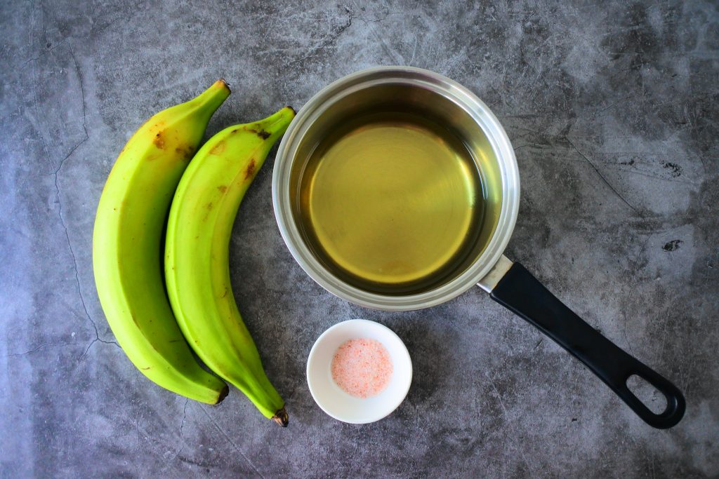 An overhead image of ingredients for fried plantain chips including green plantains, a saucepan of oil and a small dish of salt.