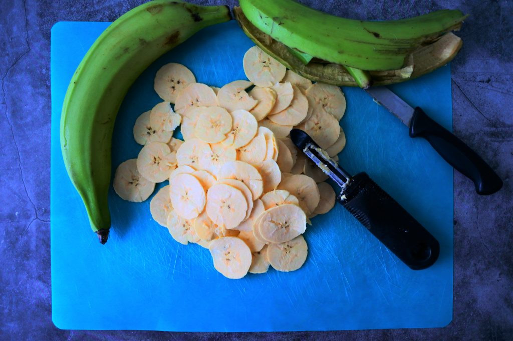 An overhead image of a cutting mat with a sliced, peeled and thin sliced rounds of a green plantain