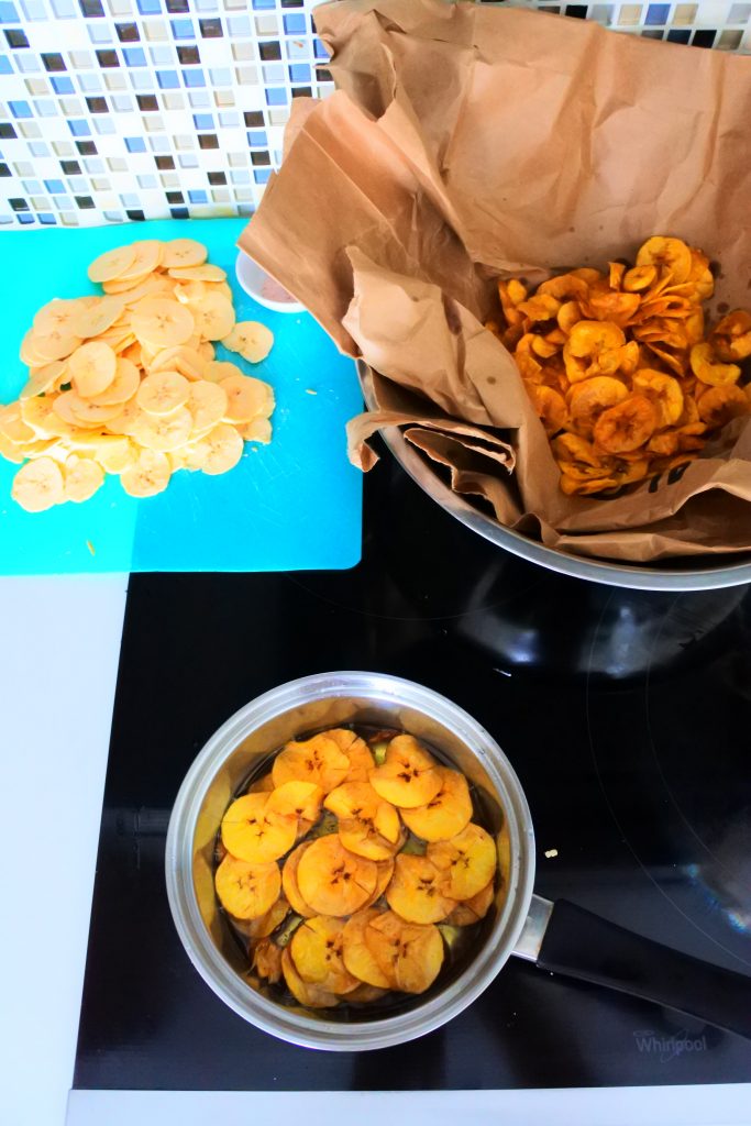 An angled overhead image of the fry station set up for plantain chips including sliced green plantains, a saucepan with oil and chips being fried and a brown paper lined bowl with already fried chips in it