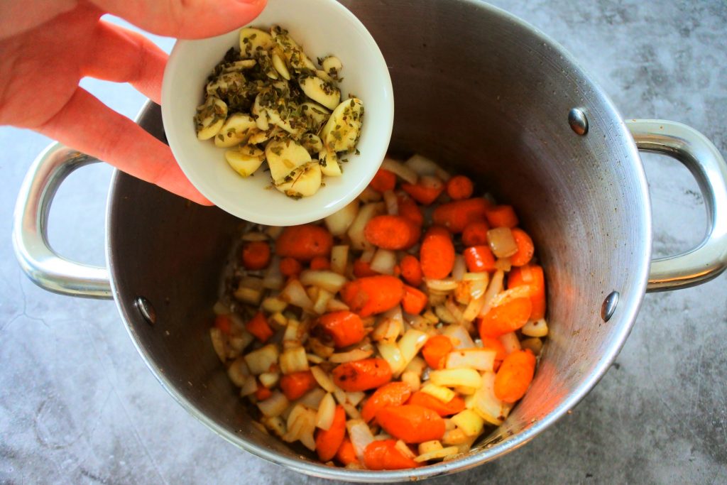 An overhead image of a pot of sautéed vegetables with sliced garlic and dried oregano being added in.