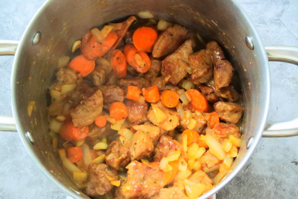 An overhead image of a pot with seared veal cubes, sautéed vegetables, gluten free flour to thicken and 1/4 cup of stock added in to deglaze the pot.