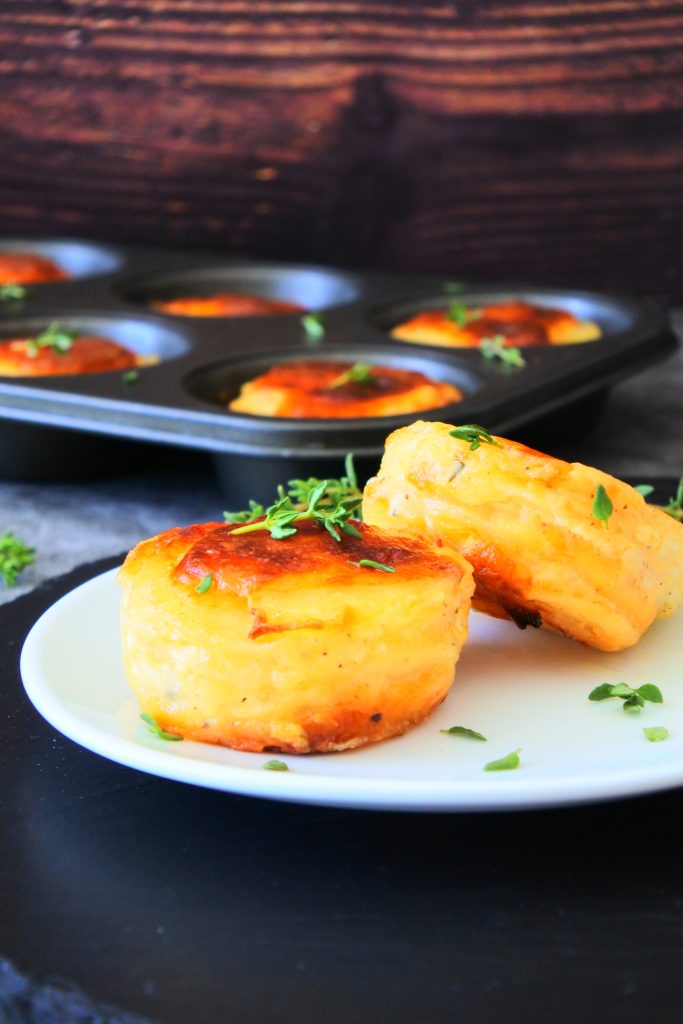 A close up image of two stacks of potato au gratin on a white plate topped with sprigs of fresh thyme with a muffin tin of other stacks in the background
