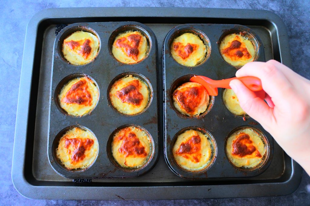 An overhead image of a baking tray with 12 freshly baked potato au gratins in muffin tins with a red silicone spatula being run around the edges to shape them before they cool and set